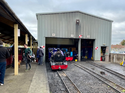 An engine shed with three tracks leading in, with numbers overhead 1/2/3. Two engines are visible -- a dark blue engine and a light blue engine. To the left-hand side you can see the carriages with doors open, and passengers milling around and ready to board.